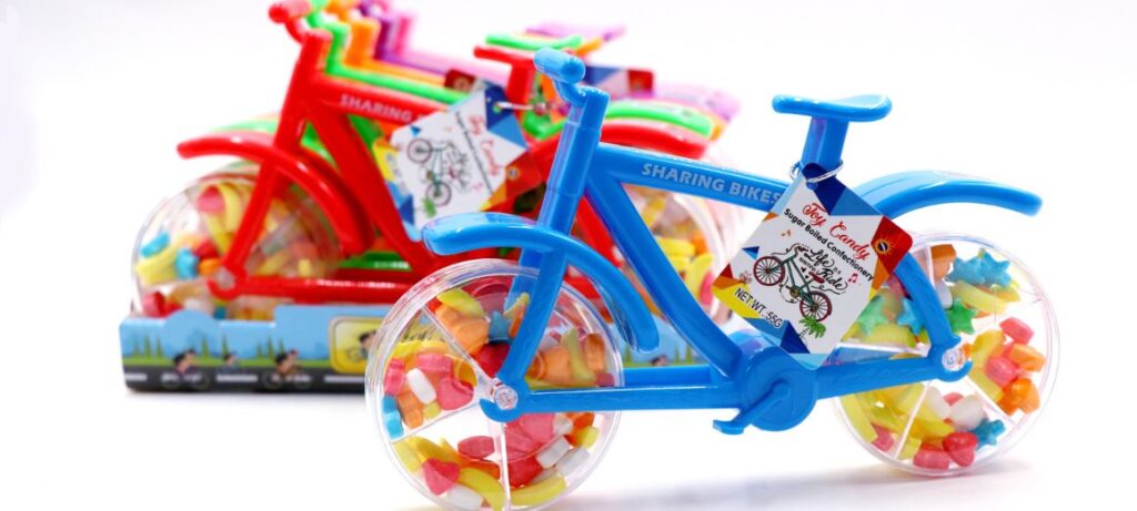 A colorful display of bicycle-shaped candy toy containers with orange, red, green, and purple handlebars, filled with multicolored pressed candy pieces, set against a two-tone yellow and blue background.