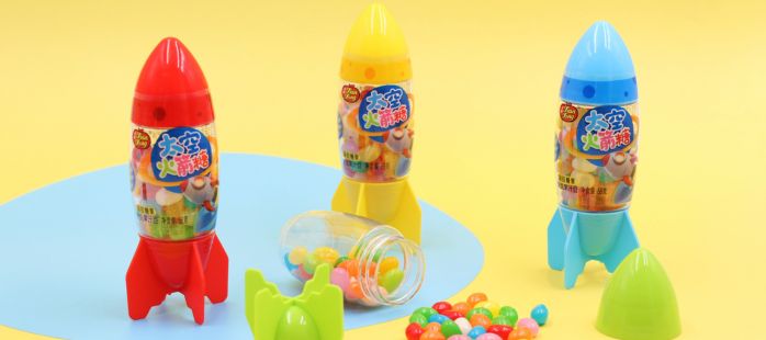 Three colorful rocket-shaped candy toy containers stand on a yellow background with a blue circle. They are red, yellow, and blue. An opened container spills multicolored jelly beans candies onto the surface.