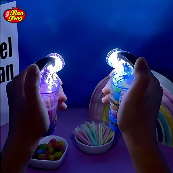 Two hands hold illuminated plastic Fire extinguisher candy toy with glowing caps against a blue background. A bowl of colorful jelly beans is on the table.