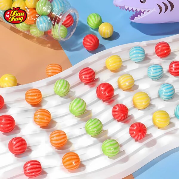 A collection of colorful, bubble gum candies scattered on a wavy white surface. They include red, green, orange, and yellow colors. A small plastic shark candy toy and a tilted jar are nearby on a multicolored background.