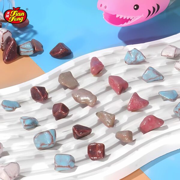 Colorful, polished stone chocolate candies are displayed on a wavy, white surface. The background features contrasting blue and brown sections. A pink inflatable shark candy toy with a smiling face is partially visible at the top right corner.