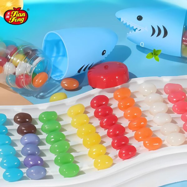 Colorful jelly beans are arranged neatly on a white tray and scattered around open shark-shaped candy toy containers. A small mint sprig is nearby,